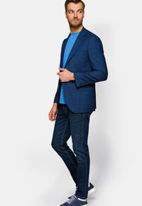 Niebieska marynarka w kratę z klapami, zapinana na jeden guzik, zestawiona z niebieską koszulką i ciemnymi dżinsami. Całość uzupełniają sportowe sneakersy.