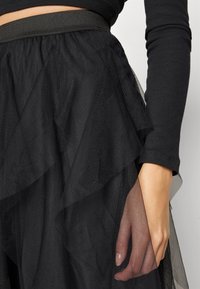 Black tulle skirt with layered ruffles, featuring a textured finish and an elastic waistband. Close-up view showing hand and fabric detail.