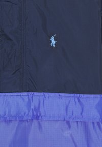 Navy and purple color-blocked nylon jacket featuring a small embroidered logo, smooth texture, and visible stitching details.