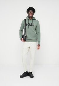 Sudadera verde con capucha y logotipo "BOSS", detalles en negro; combinada con pantalones blancos y zapatos negros; cuenta con un bolsillo frontal. Bandolera negra.