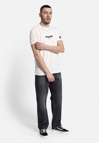 T-shirt couleur crème avec un graphique en texte noir à l'avant, associé à un jean gris foncé et des baskets noires. Design simple et décontracté.