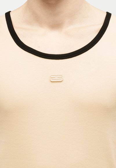 Gros plan sur un haut beige avec un col bordé de noir et un petit logo brodé centré sous la clavicule.