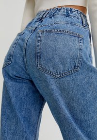 Jeans de mezclilla de color azul claro con una cintura alta y elástica, que presentan dos bolsillos traseros y costuras decorativas. La tela tiene una textura ligeramente desvaída.