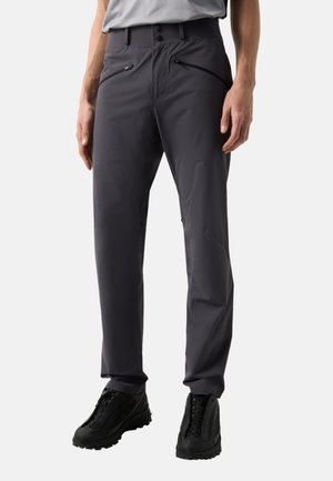 Mężczyzna noszący ciemnoszare, dopasowane spodnie z zamykanymi na zamek kieszeniami z przodu oraz czarne buty do chodzenia po górach, stojący na jasnym tle.