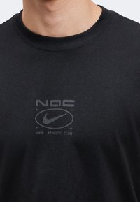Κοντινό σε μαύρο T-shirt με διακριτικό λογότυπο Nike Athletic Club και το σήμα Swoosh τυπωμένο στο στήθος.
