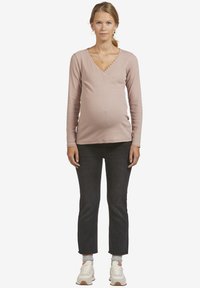 Langärmliges, pinkes Wickeloberteil mit gewelltem Ausschnitt, kombiniert mit dunklen Jeans und weißen Sneakern. Das Model ist schwanger.