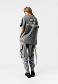 Camiseta de algodón gris de corte extragrande con "MOVIMIENTO OLA MOD" impreso en la parte posterior, combinada con pantalones cargo grises que presentan cremalleras y detalles de velcro.