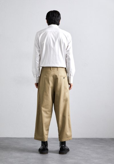 Weißes Langarmhemd und lockere khakifarbene Hose. Die Hose hat eine Gesäßtasche und umgenähte Säume. Schwarze Stiefel runden das Outfit ab.