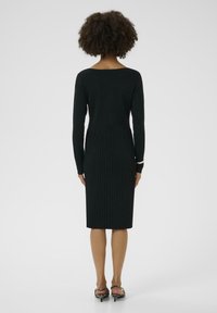 Melnas adītas kleitas ar pieguļošu augšdaļu un riboto apakšējo daļu. Garas piedurknes un izgriezums aizmugurē; saskaņota ar rakstainām kurpēm ar papēžiem.