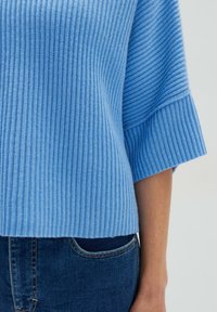 Helles blaues geripptes Sweatshirt mit kurzen Ärmeln und weiten Bündchen. Die Textur wirkt weich und wird mit dunkelblauer Jeans und einer Fronttasche kombiniert.