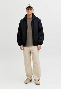 Črna lahka jakna, bež hlače, siva majica in bela baseball kapa s črnimi črtami. Čevlji so sivi z teksturirano površino.