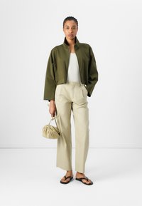 Chaqueta recortada verde oliva, camiseta blanca con textura, pantalones de pierna ancha beige, chanclas negras y un bolso beis con cordón.
