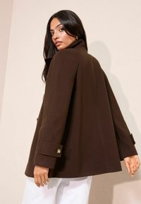 Rudas vilnos paltas su aukštu apykakle, plačiomis rankovėmis ir auksiniais mygtukais. Dizainas pasižymi tiesiu kirpimu ir lygiu audiniu.
