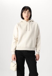 Sweat-shirt beige avec capuche et cordons, poignets et ourlet côtelés. Présente un détail de logo subtil. Assorti à un pantalon noir et un petit sac à main blanc.