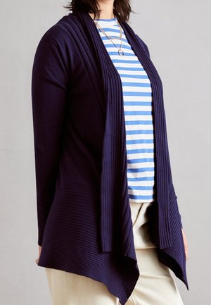 Cardigan côtelé navy à l'avant ouvert, avec une texture superposée et un ourlet drapé. Associé à une chemise à rayures bleues et blanches.