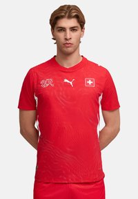 Rotes Fußballtrikot mit kurzen Ärmeln, Wellendesign, weißen Akzenten, Schweizer Flagge, Puma-Logo und Rundhalsausschnitt.