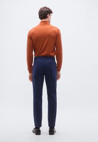 Herrtröja i figursydd modell i bränd orange med långa ärmar, kombinerad med marinblå byxor och svarta skor, sedd bakifrån.