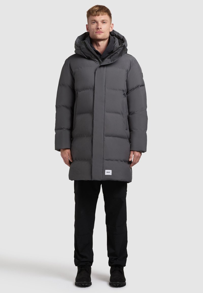 Dlhý pufrový kabát v tmavosivej farbe, s kapucňou, prešívaným dizajnom, predným zapínaním a logom pri spodnom okraji. Nosený cez čierne nohavice.