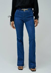 Jeans de mezclilla acampanados en azul oscuro con costuras visibles, de talle alto y acento de cinturón de cadena dorada. Textura suave y ajuste entallado.