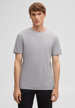 Young man with light brown hair wearing a plain grey short-sleeve crew neck t-shirt and black pants, standing against a plain background.