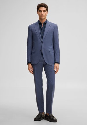 Mężczyzna stoi twarzą do przodu, ubrany w niebieski garnitur z pasującymi spodniami, ciemnoniebieską koszulę i czarne skórzane mokasyny na jasnoszarym tle.