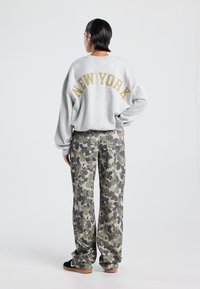 Persona con cabello oscuro corto lleva una sudadera gris con el texto "NUEVA YORK" y pantalones de camuflaje holgados, de pie contra un fondo sencillo.