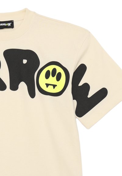 T-shirt di cotone color crema con lettering nero in grassetto e una grafica circolare gialla brillante con un volto sorridente e i canini.