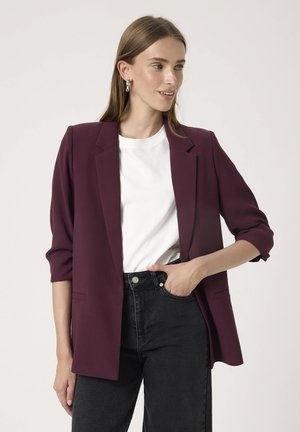 Burgunderfarbener Blazer mit strukturiertem Design, Schulterpolstern und umgeschlagenen Ärmeln, kombiniert mit einem weißen T-Shirt und hochgeschnittenen schwarzen Jeans.