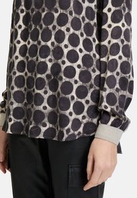 Long-sleeve shirt in light beige with large black polka dots. Features a relaxed fit and textured gray cuffs, set against black leather pants.