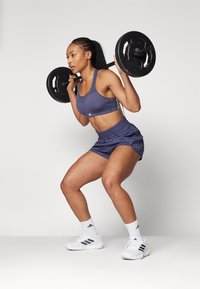 Atleta realizando una sentadilla con una barra, vistiendo un sujetador deportivo azul marino y shorts a juego. Zapatillas deportivas blancas con detalles en negro.