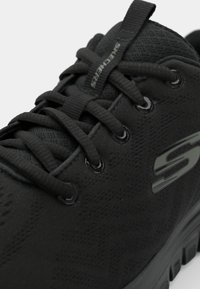 Schwarzer Sportschuh mit einer strukturierten Oberseite, runden Schnürsenkeln, Ösen und einer Gummisohle mit einem auffälligen Logo-Detail an der Seite.