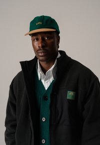 Svart fleecejacka med en grön och svart väst med ribbad textur, knappliknande detaljer och en logotypapplikation; grön keps med beige skärm.