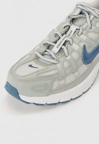 Szürke és fehér sneaker, amely hálós és szintetikus anyagokból készült, kék akcentusokkal és fűzőkkel, fényvisszaverő elemekkel és texturált talppal.
