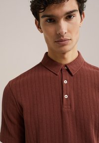 Rustfarvet polo-shirt med en struktureret overflade, korte ærmer, krave og en tre-knaps placket med hvide knapper.