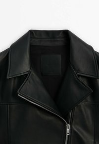 Veste en cuir noire avec un large col et une fermeture éclair argentée partiellement fermée sur le devant, sur un fond clair uni.