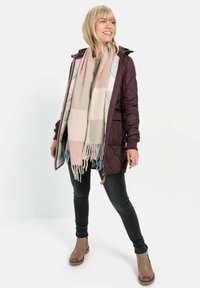 Quilted dunkelviolette Jacke mit Kapuze, getragen über einem hellen Oberteil, kombiniert mit einem gemusterten Schal in sanften Farben und blauen Akzenten. Schwarze Jeans und braune Stiefeletten sichtbar.