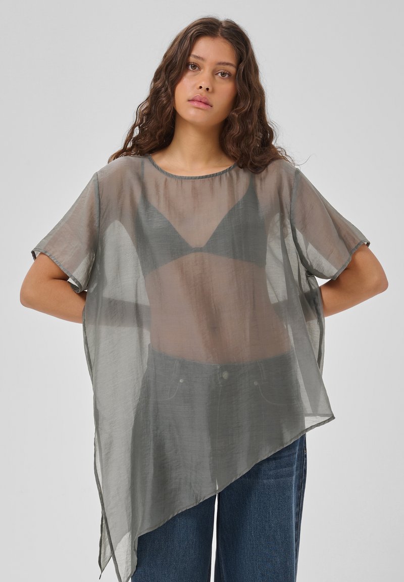 Jeune femme aux longs cheveux bouclés porte un top gris asymétrique transparent sur un bralette noir et un jean bleu, debout, les bras pliés derrière.