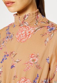 Blusa floral de tom castanho claro com colarinho alto franzido. Apresenta padrões de flores coloridos em vermelho, azul e verde contra um tecido suave e texturizado.