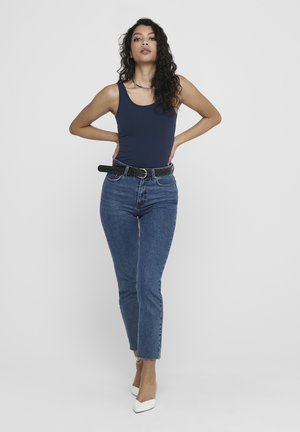 Marinefarbener Tanktop, hochgeschnittene blaue Jeans mit leichtem Verblassen, schwarzer gepunkteter Gürtel und weiße spitze Absätze. Lockige Haare, stehende Pose.