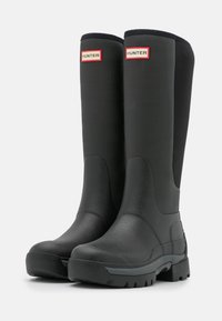 Mustat kumiset sateenvarvastossut, joissa on karkea pinta, hieman kaareva varsi ja kestävä uritettu pohja. Varustettu punaisilla logo-aksenteilla.