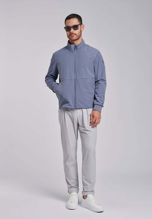 Mężczyzna w okularach przeciwsłonecznych, niebieskiej kurtce zapinanej na zamek, jasnoszarych spodniach i białych trampkach, stojący z jedną ręką w kieszeni na jednolitym tle.