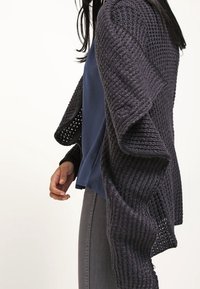 Marinblå stickad cardigan med öppen fram, strukturerat mönster och voluminösa ärmar, bärs över en blå topp och matchas med grå jeans.