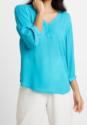 Woman wearing a loose turquoise long-sleeve blouse with rolled sleeves and white pants, hand touching neck, against a plain background.