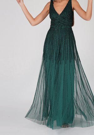 Mujer con un vestido formal sin mangas de color verde oscuro con adornos de cuentas verticales y una falda transparente y fluida, de pie con los brazos levantados.