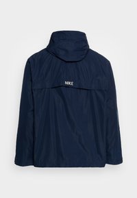 Granatowa kurtka Nike z kapturem, o luźnym kroju, gładkiej teksturze i białym logo na plecach. Prosty design bez widocznych zapięć.