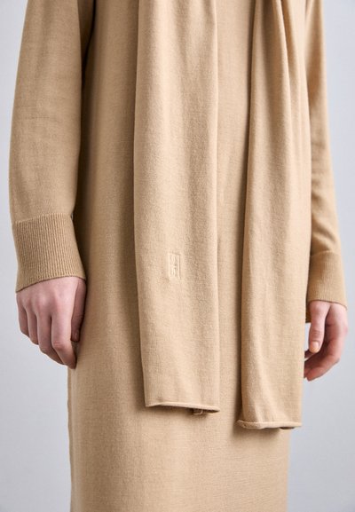 Beige strikket kjole med lange ærmer og ribbede manchetter, med et matchende tørklæde draperet foran. Subtil logo detalje på kjolen.