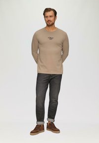 Beige langarmshirt met logo op de borst, gecombineerd met donkere jeans en bruine sneakers, wat een casual maar stijlvolle uitstraling laat zien.