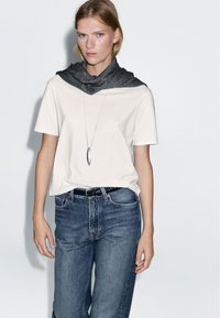 Kortärmad beige t-shirt, mörkgrå scarf draperad, blå denimjeans, svart flätad bälte, silverhänge, enkel design, avslappnad stil.