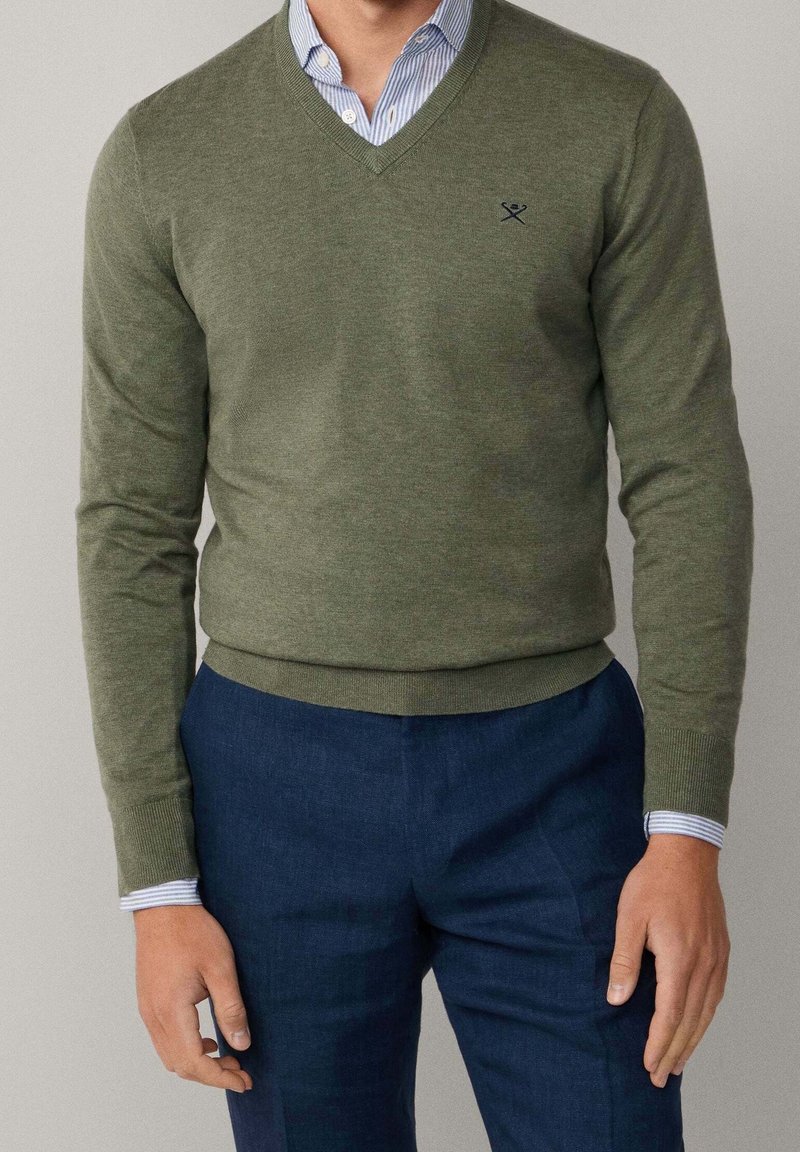 Homme portant un pull col V vert olive par-dessus une chemise rayée bleue, accompagné d’un pantalon bleu foncé, debout, les mains détendues le long du corps.