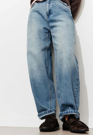 Personne portant un jean bleu clair à jambes larges avec des revers roulés et des mocassins foncés à motifs devant un mur blanc uni.